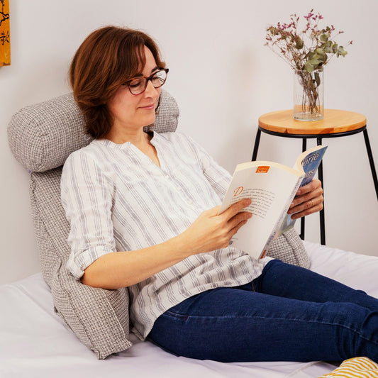 Femme qui lit un livre au lit, adossée à un coussin de lecture gris clair