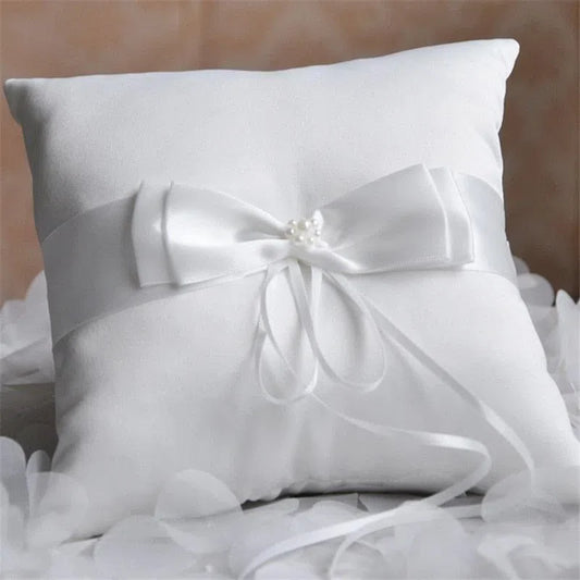 Coussin porte-alliances blanc orné d'un nœud de satin, posé sur du tulle et des pétales blancs