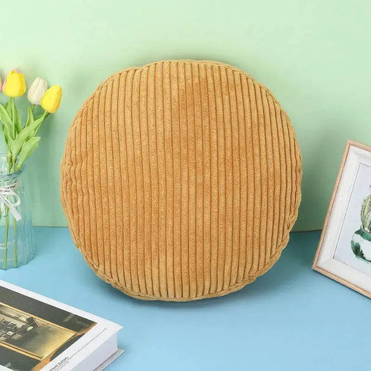 Coussin rond en velours côtelé couleur ocre posé debout contre un mur vert, à côté d'un vase de tulipes