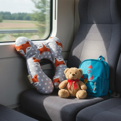 Coussin de voyage enfant en U à motif renard posé sur un siège de train, avec un sac à dos et un ourson