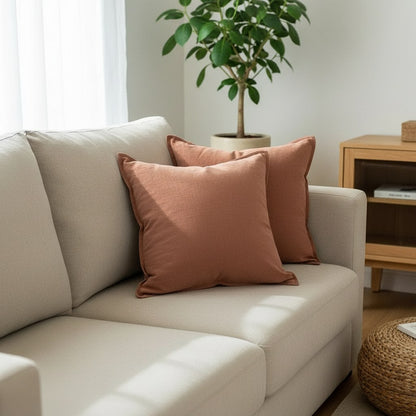 Coussins terracotta à bord plat, aspect toile lavée, sur un canapé blanc avec un pouf en rotin